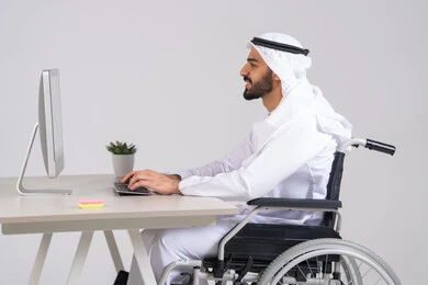 Executing daily tasks, managing and organizing business affairs through technical devices, integrating work with technologies and technology, administrative and office job professions, a side view of an Emirati Gulf Arab man wearing a kandura and ghutrah sitting in a wheelchair working on a computer inside the office.