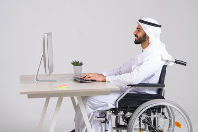 إنجاز المهام العمل المطلوبة، إدارة شؤون الأعمال وتنظيمها عبر الاجهزة التقنية، دمج العمل مع التقنيات والتكنولوجيا، مهن ووظائف ادارية مكتبية, صورة من الجانب لرجل عربي خليجي اماراتي يرتدي الكندورة و الغترة يجلس على كرسي متحرك يعمل على الحاسوب داخل المكتب