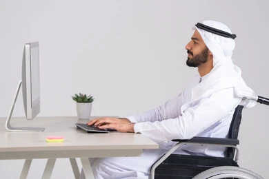 إدارة شؤون الأعمال وتنظيمها عبر الاجهزة التقنية، دمج العمل مع التقنيات والتكنولوجيا، مهن ووظائف ادارية مكتبية, صورة من الجانب لرجل عربي خليجي اماراتي يرتدي الكندورة و الغترة يجلس على كرسي متحرك يعمل على الحاسوب داخل المكتب، إنجاز المهام العمل المطلوبة