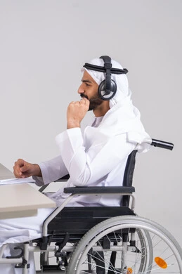 Providing answers to customer questions, communicating with customers to resolve issues and complaints, using headphones and advanced technologies at work, a side image of an Emirati Gulf Arab man wearing a kandura and ghutrah sitting in a wheelchair working in customer service.