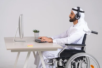 التواصل مع الزبائن لحل المشكلات والشكاوي، استخدام الهيدفون والتقنيات المتطورة في العمل، الاجابه على اسئلة العملاء، صورة من الجانب لرجل عربي خليجي اماراتي يرتدي الكندورة و الغترة يجلس على كرسي متحرك يعمل  فى مجال خدمة العملاء ، اجراء اتصال من خلال جهاز الحاسوب