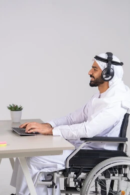 استخدام الهيدفون والتقنيات المتطورة في العمل، التواصل مع الزبائن لحل المشكلات والشكاوي، الاجابه على اسئلة العملاء، صورة من الجانب لرجل عربي خليجي اماراتي يرتدي الكندورة و الغترة يجلس على كرسي متحرك يعمل  فى مجال خدمة العملاء ، اجراء اتصال من خلال جهاز الحاسوب