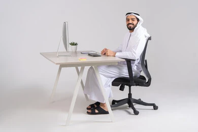 Looking at the camera with gestures of happiness and joy, using modern and advanced technological devices, following up and organizing administrative work, an Emirati Gulf Arab employee wearing a kandura and ghutrah sitting behind a wooden table with a computer in front of him.