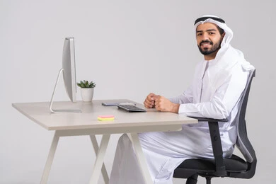The use of modern and advanced technological devices, monitoring and organizing administrative work, an Arab Gulf Emirati employee wearing a kandura and ghutrah sitting behind a wooden table with a computer in front of him, looking at the camera with gestures of happiness and joy.