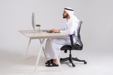 Completing work tasks in the office, the development of Emirati companies, managing and organizing business affairs, a side view of an Arab Gulf Emirati employee sitting on a comfortable chair wearing a kandura and ghutrah working on a computer.