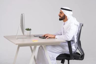 تطور الشركات الاماراتية، إتمام مهام العمل في المكتب، إدارة شؤون الأعمال وتنظيمها،, صورة من الجانب  لموظف عربي خليجي اماراتي يجلس على كرسي مريح يرتدي الكندورة و الغترة يعمل  على جهاز الحاسوب