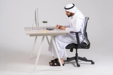 كتابة وتسجيل الملاحظات والنتائج, صورة من الجانب لموظف عربي خليجي اماراتي يرتدي الكندورة و الغتره يجلس امام الحاسوب يقوم بإنجاز المهام اليومية، دمج الأعمال مع التقنية و التكنولوجيا, تحقيق أهداف الشركة