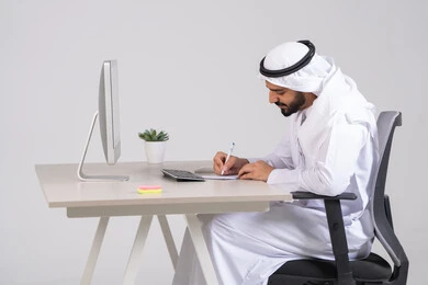 تدوين و تسجيل النتائج و الملاحظات, صورة من الجانب لموظف عربي خليجي اماراتي يرتدي الكندورة و الغتره يجلس امام الحاسوب يقوم بإنجاز المهام اليومية، دمج الأعمال مع التقنية و التكنولوجيا, تحقيق أهداف الشركة