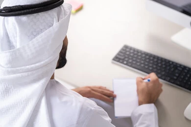 حفظ وتدوين الشكاوى والملاحظات، التخطيط اليومي وإدارة الوقت، ادارة المهام وتنظيم الأعمال،, صورة من الخلف لشاب عربي خليجي اماراتي يرتدي الثوب التقليدي يقوم بكتابة الأهداف اليومية
