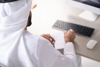 Recording and documenting notes and complaints, daily planning and time management, task management and organizing work, a picture from behind of a young Arab Gulf Emirati man wearing traditional attire writing daily goals.