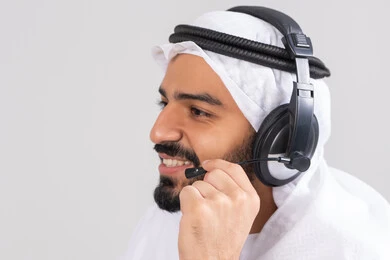 Answering and responding to inquiries and questions, using headphones and advanced technologies at work, communicating with customers to solve problems and complaints, a close-up side image of a young Emirati Gulf Arab man wearing a kandura and ghutrah, wearing headphones, working in the customer service department.