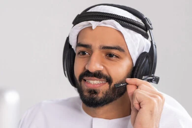 استخدام سماعات الرأس والتقنيات المتطورة في العمل، التواصل مع الزبائن لحل المشكلات والشكاوي، الإجابة والرد على الاستفسارات والاستعلامات, صورة مقربة لشاب عربي خليجي اماراتي يرتدي الكندورة و الغتره يضع الهيدفون يعمل في قسم خدمة العملاء