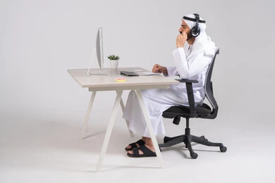Working on computers at the company headquarters, answering questions and inquiries, a side image of a young Arab Gulf Emirati man wearing a kandura and ghutrah, wearing headphones, communicating with customers to resolve issues and complaints, integrating business with technology and technology.