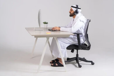 إدارة العمليات وتنظيم الأعمال، الإجابة على الأسئلة والاستفسارات، مفهوم خدمة العملاء، صورة من الجانب لموظف عربي خليجي اماراتي يرتدي الكندورة و الغتره و سماعات الرأس يعمل على الحاسوب في المكتب، النقر على لوحة المفاتيح
