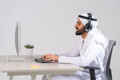 Organizing and managing the company's business, answering questions and inquiries, the concept of customer service, a side view of an Emirati Gulf Arab employee wearing a kandura and ghutrah, with headphones, working on a computer in the office, typing on the keyboard.