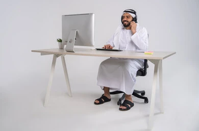 Management and organization of the company's work, integrating work with technology and technology, office professions and jobs, inquiries and communication department, answering questions and inquiries, the concept of customer service, an Emirati Gulf Arab employee wearing a kandura and ghutrah and headphones working on a computer in the office, typing on the keyboard.