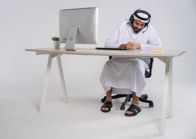 The concept of customer service, following up and developing work, responding to inquiries and questions, recording and documenting notes and complaints, an Emirati Gulf Arab employee wearing a kandura and ghutrah sitting in front of the computer with headphones on his head writing on papers.