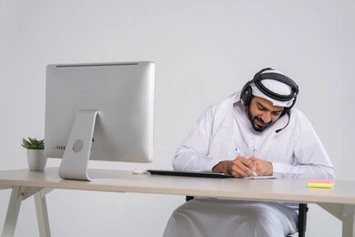Monitoring and developing work, responding to inquiries and questions, recording and documenting notes and complaints, an Emirati Gulf Arab employee wearing a kandura and ghutrah sits in front of the computer with headphones on his head writing on papers, the concept of customer service.