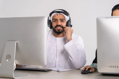 الاجابه على اسئلة العملاء، موظف عربي خليجي اماراتي يرتدي الكندورة و الغتره يرتدي سماعة الاتصال، استعلامات واستفسارات، اجراء اتصال فيديو من خلال الحاسوب ، متابعة وتطوير العمل