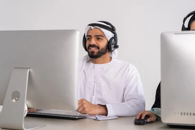 متابعة وتطوير العمل، الاجابه على اسئلة العملاء، موظف عربي خليجي اماراتي يرتدي الكندورة و الغتره يرتدي سماعة الاتصال، استعلامات واستفسارات، اجراء اتصال فيديو من خلال الحاسوب