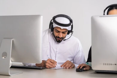 الرد على الاستفسارت والتساؤولات، تدوين وتسجيل الملاحظات والشكاوي، موظف عربي خليجي اماراتي يرتدي الكندورة و الغتره يجلس امام الحاسوب يضع سماعات الهيدفون على رأسه يقوم بالكتابة على الاوراق، متابعة وتطوير العمل، مفهوم خدمة العملاء