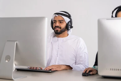 ايماءات الاندماج و التركيز، دمج التقنيات المتطورة مع العمل, التواصل مع الزبائن لحل المشكلات في مكاتب خدمة العملاء، الاجابة على الاستفسارات والاستعلامات, رجل عربي خليجي إماراتي يرتدي الكندورة والغترة يضع الهيدفون يجلس امام الحاسوب ، النقر على لوحة المفاتيح