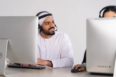 التواصل مع الزبائن لحل المشكلات في مكاتب خدمة العملاء، دمج التقنيات المتطورة مع العمل، الاجابة على الاستفسارات والاستعلامات, رجل عربي خليجي إماراتي يرتدي الكندورة والغترة يضع الهيدفون يجلس امام الحاسوب و ينظر الى شيء ما