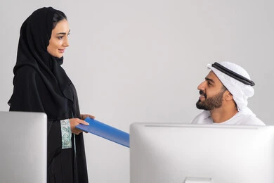 Organizing daily tasks, completing office work, an Arab Gulf Emirati woman wearing an abaya and hijab handing a blue file to her colleague, collecting data and solving problems, developing and studying new projects in companies in the United Arab Emirates.