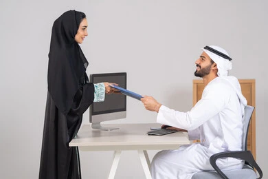 Completing office tasks, following up on daily tasks, an Arab Gulf Emirati woman wearing an abaya and hijab is handing a blue file to her colleague, collecting data and solving problems, developing and studying new projects in companies in the United Arab Emirates.