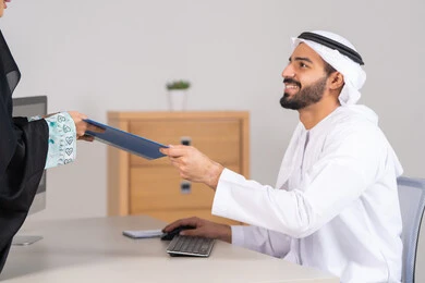 Monitoring daily tasks, completing office work, an Arab Gulf Emirati woman wearing an abaya and hijab handing over a blue file to her colleague, collecting data and solving problems, developing and studying new projects in companies in the United Arab Emirates.