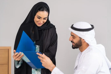 تطوير ودراسة المشاريع الجديدة في شركات الامارات العربية المتحدة، جمع البيانات وحل المشكلات، اجتماع ومناقشة عمل في مقر الشركة، رجل أعمال عربي خليجي اماراتي يرتدي الكندورة والغترة يقوم بامساك ملف عمل ازرق  يقوم بالمناقشة مع زميلته داخل المكتب