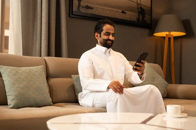 Gestures of happiness and joy, browsing the internet on a mobile phone, a Gulf Arab Saudi man wearing a white thobe sitting in the living room using his mobile phone.