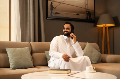 اجراء مكالمة هاتفية عبر الهاتف المحمول ، رجل عربي خليجي سعودي يرتدي الثوب الابيض يجلس فى غرفه المعيشة يستخدم الجوال، ايماءات السعادة و السرور