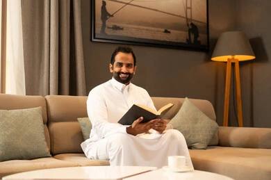 The culture of self-learning, harmony, calmness, and tranquility, enjoying reading a book, an Arab Gulf Saudi man wearing a white thobe sitting in the living room holding a book in his hand, gestures of happiness and joy, spending enjoyable time. The culture of self-learning, harmony, calmness, and tranquility, enjoying reading a book, an Arab Gulf Saudi man wearing a white thobe sitting in the living room holding a book in his hand, gestures of happiness and joy, spending enjoyable time.