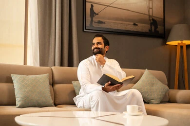 Looking and thinking about something, the culture of self-learning, harmony, calmness, and tranquility, enjoying reading a book, an Arab Gulf Saudi man wearing a white thobe sitting in the living room holding a book in his hand, gestures of happiness and joy, spending enjoyable time.