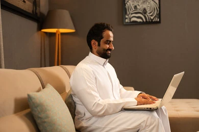 إنجاز مهام العمل عن بعد، إدارة شؤون الأعمال وتنظيمها عبر اللابتوب، ايماءات السعادة والسرور، استخدام جهاز تقني حديث ومتطور، رجل عربي خليجي سعودي يرتدي الثوب الابيض يجلس فى غرفه المعيشة يستخدم الحاسوب المحمول