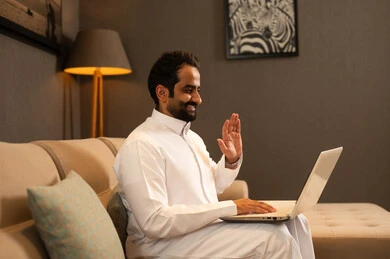 اجراء مكالمة فيديو، رفع اليد بتعابير التحية و السلام، إنجاز مهام العمل عن بعد، ايماءات السعادة والسرور، استخدام جهاز تقني حديث ومتطور، رجل عربي خليجي سعودي يرتدي الثوب الابيض يجلس فى غرفه المعيشة يستخدم الحاسوب المحمول