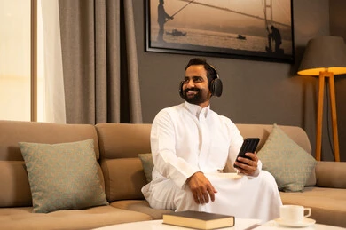 الاستفادة من الجوال والأجهزة التكنولوجية، الاستماع إلى الموسيقى والتسجيلات الصوتية، رجل عربي خليجي سعودي يرتدي الثوب الابيض يجلس فى غرفه المعيشة يرتدي سماعة الرأس بإيماءات السعادة, استخدام الهاتف و تقنية الهيدفون