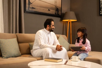 Expressions of happiness and joy, a Gulf Arab Saudi girl wearing casual clothes sitting in the living room using a tablet alongside her father, following an online educational platform, integrating learning with technology, parents' interest in their children, watching a recreational and educational activity.