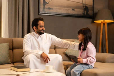 فتاة عربية خليجية سعودية ترتدي لباس الكاجوال تجلس فى غرفه المعيشة تستخدم التابلت تجلس برفقة والدها، متابعة منصة التعليم الالكتروني عبر الانترنت، دمج التعلم بالتكنولوجيا، اهتمام الآباء بالأبناء، مشاهدة نشاط ترفيهي و تعليمي، تعابير السعادة و السرور