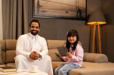 متابعة منصة التعليم الالكتروني عبر الانترنت، دمج التعلم بالتكنولوجيا، اهتمام الآباء بالأبناء، مشاهدة نشاط ترفيهي و تعليمي، فتاة عربية خليجية سعودية ترتدي لباس الكاجوال تجلس فى غرفه المعيشة تستخدم التابلت تجلس برفقة والدها، تعابير السعادة و السرور