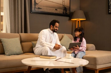 Integrating learning with technology, parents' interest in their children, watching a recreational and educational activity, following online educational platforms, an Arab Gulf Saudi girl wearing casual clothes sitting in the living room using a tablet alongside her father, expressions of happiness and joy.