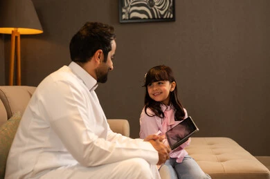 مشاهدة نشاط ترفيهي و تعليمي، اهتمام الآباء بالأبناء، دمج التعلم بالتكنولوجيا، متابعة منصات التعليم الالكتروني عبر الانترنت، فتاة عربية خليجية سعودية ترتدي لباس الكاجوال تجلس فى غرفه المعيشة تستخدم التابلت تجلس برفقة والدها، تعابير السعادة و السرور