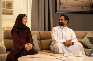 الالفة والمحبة بين الزوجين، اجواء عائلية سعيدة, مفهوم التحدث ومناقشة الاخبار, رجل عربي خليجي سعودي يرتدي الثوب الابيض يجلس فى غرفه المعيشة برفقة زوجته, ايماءات السرور و الفرح