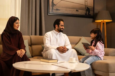 Spending happy family time, integrating learning with technology, following online e-learning platforms, a Gulf Arab Saudi girl wearing casual clothes sitting in the living room using a tablet alongside her parents, expressions of happiness and joy.