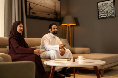 The concept of entertainment and leisure, cultural programs and news, watching movies and series, remote control using modern electronic devices, an Arab Gulf Saudi man wearing a white thobe sitting in the living room with his wife holding the remote control in his hands.