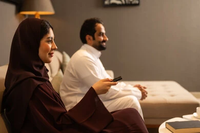 Watching movies and series, controlling modern electronic devices remotely, an Arab Gulf Saudi woman wearing a hijab and an abaya sitting in the living room with her husband holding a remote control in her hand, cultural programs and news.