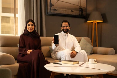 قضاء وقت عائلي ممتعه، استخدام الاجهزة التقنية، عرض شاشة هاتف محمول سوداء فارغة، ؤجل عربي خليجي سعودي يرتدي الثوب الابيض يجلس فى غرفه المعيشة برفقة زوجته يمسك بيدية الجوال ، تعابير السعادة و السرور
