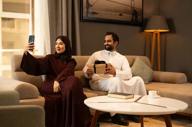 Taking a selfie with a mobile phone, documenting happy moments, sharing and giving gifts during holidays and family occasions, an Arab Gulf Saudi man wearing a white thobe sitting in the living room with his wife holding a gift in his hands to celebrate their wedding anniversary, the concept of family bonding.