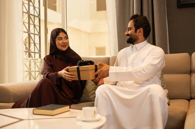 Sharing and giving gifts during holidays and family occasions, a Saudi Arabian Gulf man wearing a white thobe sits in the living room presenting a gift to his wife on the occasion of their wedding anniversary, the concept of family bonding, expressions and gestures of happiness and joy, intimacy and love between the spouses.